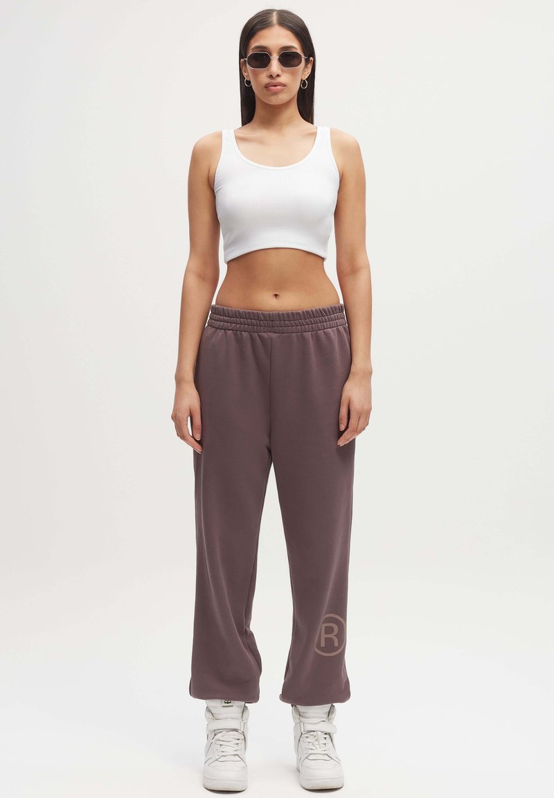 Witte geribbelde crop top en losvallende, grijsbruine joggingbroek met een logo. Draagt witte hoge sneakers. Gladde stofstructuur.