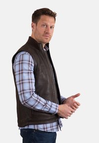 Brown leather vest with a high collar and front zipper, worn over a blue and white plaid shirt. Smooth texture, fitted design, no sleeves.