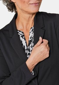 Zwarte blazer met een notch-kraag, gedragen over een gestreepte zwart-wit blouse. De textuur is glad en de pasvorm lijkt op maat gemaakt.