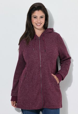 Vrouw met lang donker haar die een gestructureerde paarse zip-up hoodie met zakken draagt, glimlachend, staand tegen een effen witte achtergrond.