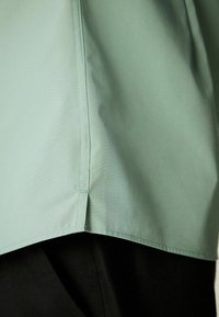 Jasnozielona bluzka o gładkiej fakturze, z prostym dołem i czarnym materiałem poniżej. Wyróżnia się starannymi szwami i minimalistycznym wzornictwem.