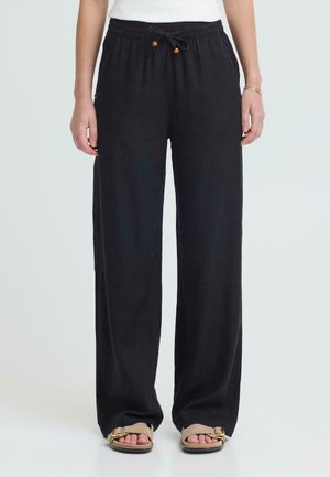 Osoba nosząca czarne spodnie z szerokimi nogawkami, z elastycznym paskiem i sznurkiem do wiązania, w połączeniu z beżowymi sandałami z odkrytymi palcami i białym topem.