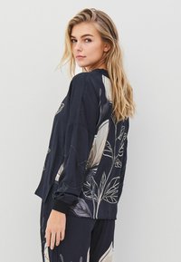 Zwart shirt met lange mouwen van lichtgewicht stof, featuring een bloemenpatroon in beige en lichtgrijs op de achterkant en mouwen. Gelaagd afwerking.