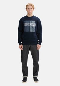 Sudadera azul marino con un estampado gráfico en el centro, combinada con jeans oscuros y zapatos. Ajuste casual, puños y cuello de canalé.
