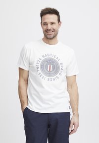 Balta medvilninė marškinė su apvaliu logotipo dizainu, turinčiu tamsiai mėlyną tekstą ir detales. Trumpi rankoviai ir trumpa apykaklė, derinama su tamsiai mėlynomis kelnėmis.