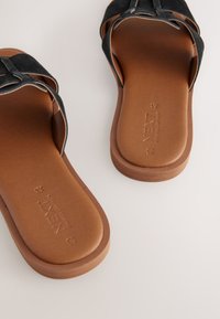 Sandales slides en daim noir avec une semelle plate en caoutchouc marron. Elles comportent deux brides superposées et un logo de la marque embossé sur la semelle intérieure.