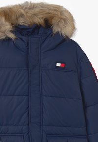 Námořnická modrá péřová bunda s kapucí lemovanou kožešinou a malou obdélníkovou nášivkou s červenou, bílou a modrou na hrudi.