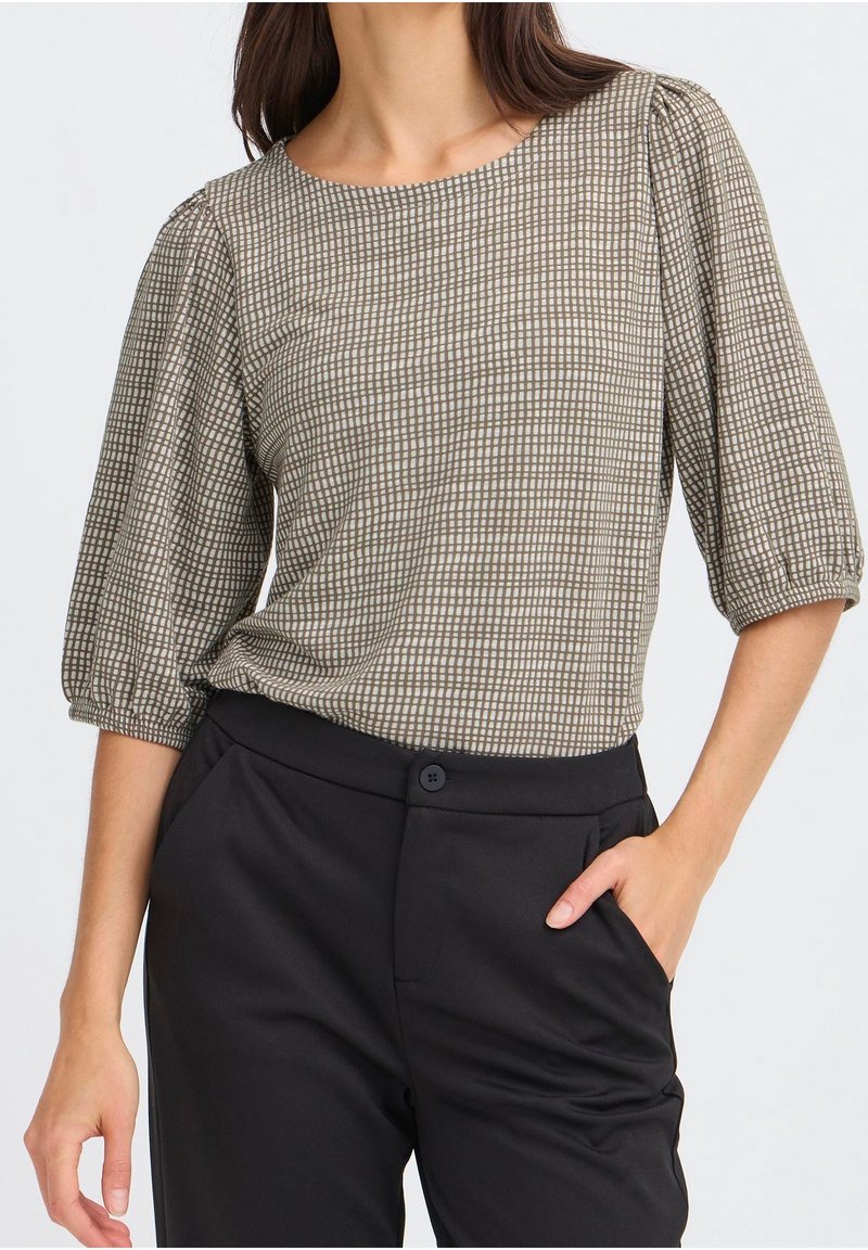 Femme portant un chemisier beige à carreaux avec des manches bouffantes et un pantalon noir taille haute, la main dans une poche.