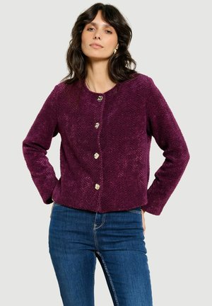 Frau mit dunklen Haaren, die eine lila strukturierte, geknöpfte Jacke und blaue Jeans trägt, steht mit den Händen in den Hüften vor einem einfarbigen Hintergrund.