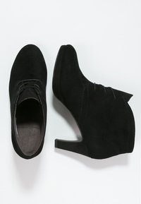 Botas de tobillo de ante negro con tacón cónico, que presentan un diseño de cordones y una textura suave. La suela es plana y ligeramente curva.