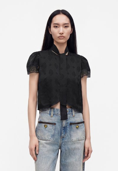 Femme aux longs cheveux noirs porte un chemisier noir à manches courtes avec poignets en dentelle et un jean bleu clair avec grandes poches avant et détails de boutons.