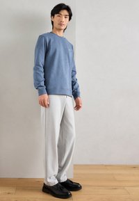 Sweatshirt de algodão azul claro com punhos canelados, combinado com calças cinzentas claras e sapatos pretos, em pé sobre um chão de madeira contra uma parede neutra.