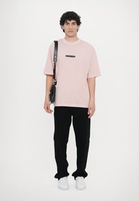 Camiseta de algodão rosa com um patch de logotipo preto, combinada com calças pretas e ténis brancos. Acessórios incluem uma mala de ombro preta.