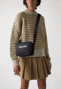 Borsa a tracolla nera con una texture liscia, hardware in argento e un dettaglio con logo. Indossata con un maglione a righe in maglia e una gonna plissettata.