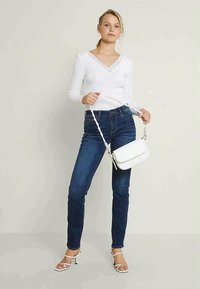 Λευκό μακρυμάνικο μπλουζάκι με δαντελένιες λεπτομέρειες, μπλε στενά τζιν, λευκή τσάντα χειρός με πλεχτό λουρί και λευκά ψηλοτάκουνα σανδάλια με λουράκια.