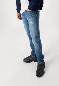 Vaqueros de mezclilla de color azul claro con detalles desgastados, combinados con zapatillas negras texturizadas. Los vaqueros presentan un corte recto y un ligero desvanecimiento.