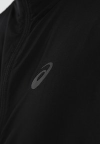 Schwarze Sportjacke mit hohem Kragen, ausgestattet mit einer glatten Textur und einem grauen Logo auf der linken Seite. Sichtbarer Reißverschluss.