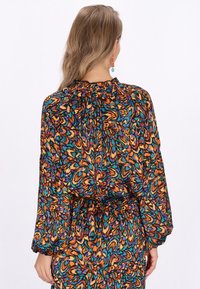 Blusa colorata e patterned in arancione, verde teal, viola e nero. Presenta un colletto arricciato e maniche lunghe e vaporose. Texture del tessuto morbida.