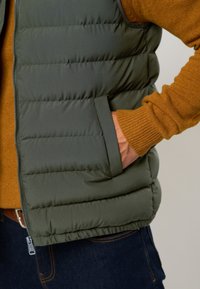 Vest verde puf din material cu cusături orizontale, cu fermoar în față și un buzunar lateral; purtat peste un pulover muștar, cu manșete ribbate.