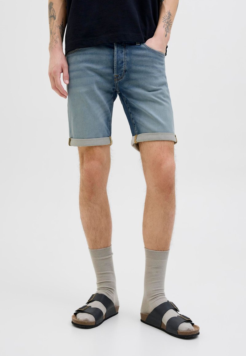 Man die opgerolde denimshort draagt, lichtgrijze sokken en zwarte sandalen, staand met één hand in de zak tegen een witte achtergrond.