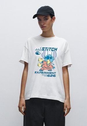 Person wearing a white T-shirt with a colorful Stitch character from Lilo & Stitch, text "STITCH EXPERIMENT 626," and black pants and cap.