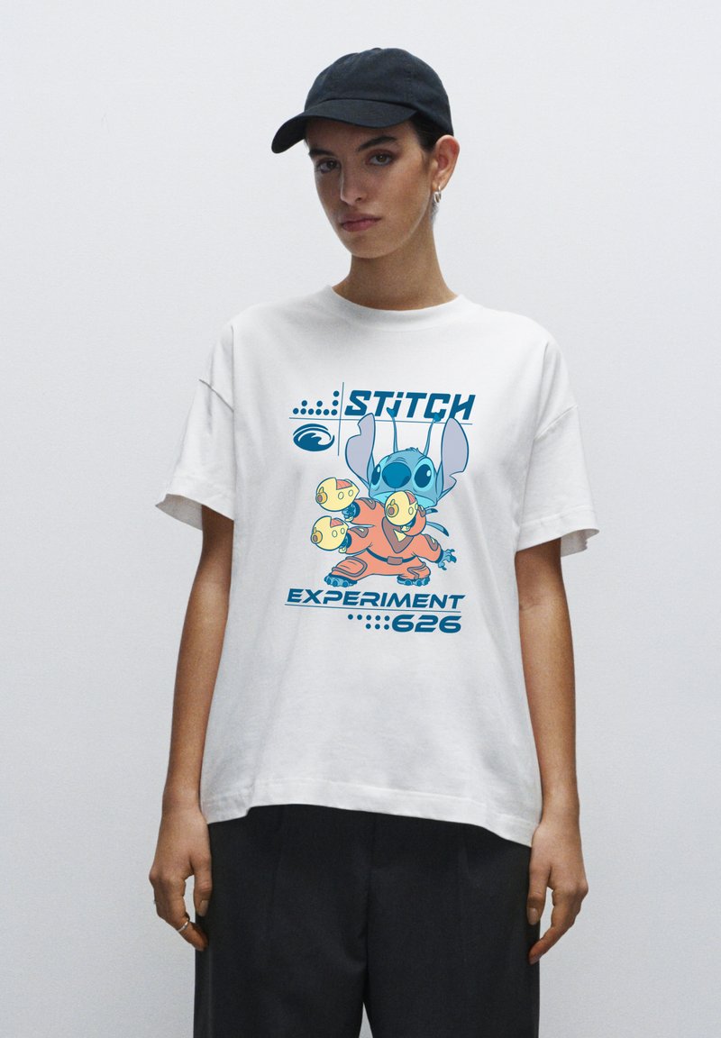 Osoba v bílém tričku s barevným charakterem Stitch z filmu Lilo a Stitch, s textem "STITCH EXPERIMENT 626" a v černých kalhotách a kšiltovce.