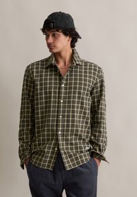 Jongen draagt een groene geruite blouse, donkere corduroy broek, zwarte pet achterstevoren en een zilveren ketting, met zijn handen in zijn zakken.