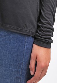 Hand resting by side wearing dark long-sleeve top and blue denim jeans, showing sleeve wrinkles and small embroidered logo on shirt hem.