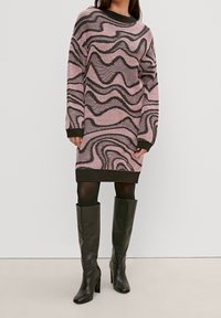 Robe en tricot rose avec des motifs ondulés noirs, encolure côtelée et poignets. Associée à des bottes en cuir noir jusqu'aux genoux.