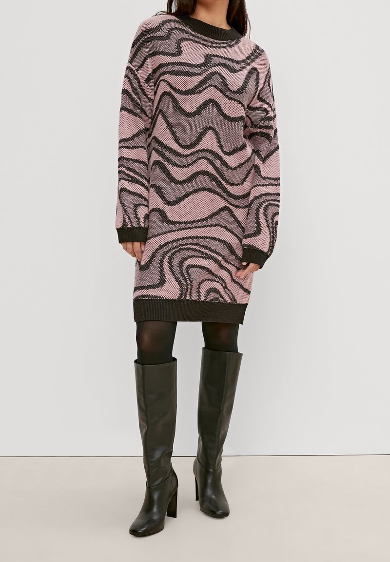 Robe en tricot rose avec des motifs ondulés noirs, encolure côtelée et poignets. Associée à des bottes en cuir noir jusqu'aux genoux.