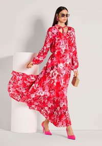 Maxikleid in leuchtendem Pink mit rotem Blumenmuster, langen Ärmeln, Bindeband am Hals und ausgestellt geschnittenem Saum, kombiniert mit pinken High Heels und geflochtener Handtasche.