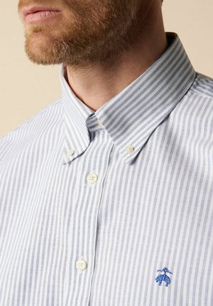 Brodaty mężczyzna ubrany w jasnoniebieską i białą koszulę zapinaną na guziki z pionowymi paskami oraz małym niebieskim wyszywanym logo na piersi.