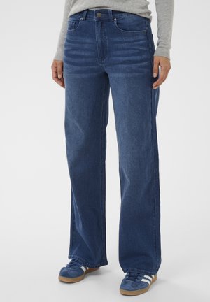 Frau trägt hochgeschnittene, weitgeschnittene blaue Jeans, ein graues Langarmshirt und blaue Turnschuhe mit weißen Streifen vor einem schlichten Hintergrund.