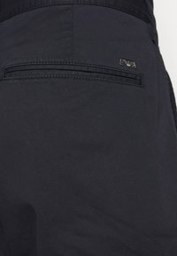 Mörkblå byxor med en slät textur, med en bakficka med slits och en metallisk logoprydnad. Klassisk och figursydd design.