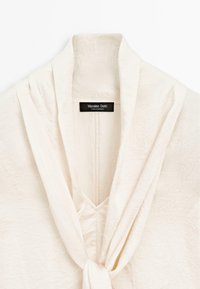Blusa beige chiaro, con texture, dotata di scollo a V e fronte drappeggiato. Presenta un'etichetta con la dicitura "Massimo Dutti" nella zona del colletto.