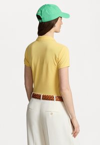 Žluté polo tričko s krátkými rukávy, nošené s krémovými kalhotami a pleteným hnědým páskem. Na fotografii je také vidět zelená baseballová čepice.