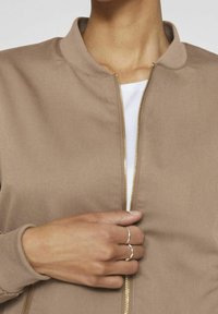 Personne fermant une veste beige sur une chemise blanche, portant deux bagues en argent sur le majeur.