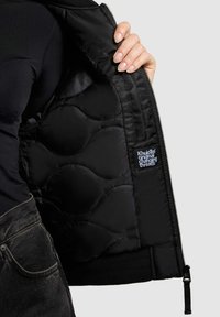 Schwarze gesteppte Jacke mit sichtbarer Polsterung im Inneren, Reißverschluss und geripptem Saum. Verfügt über ein Logotag auf dem Innenfutter.