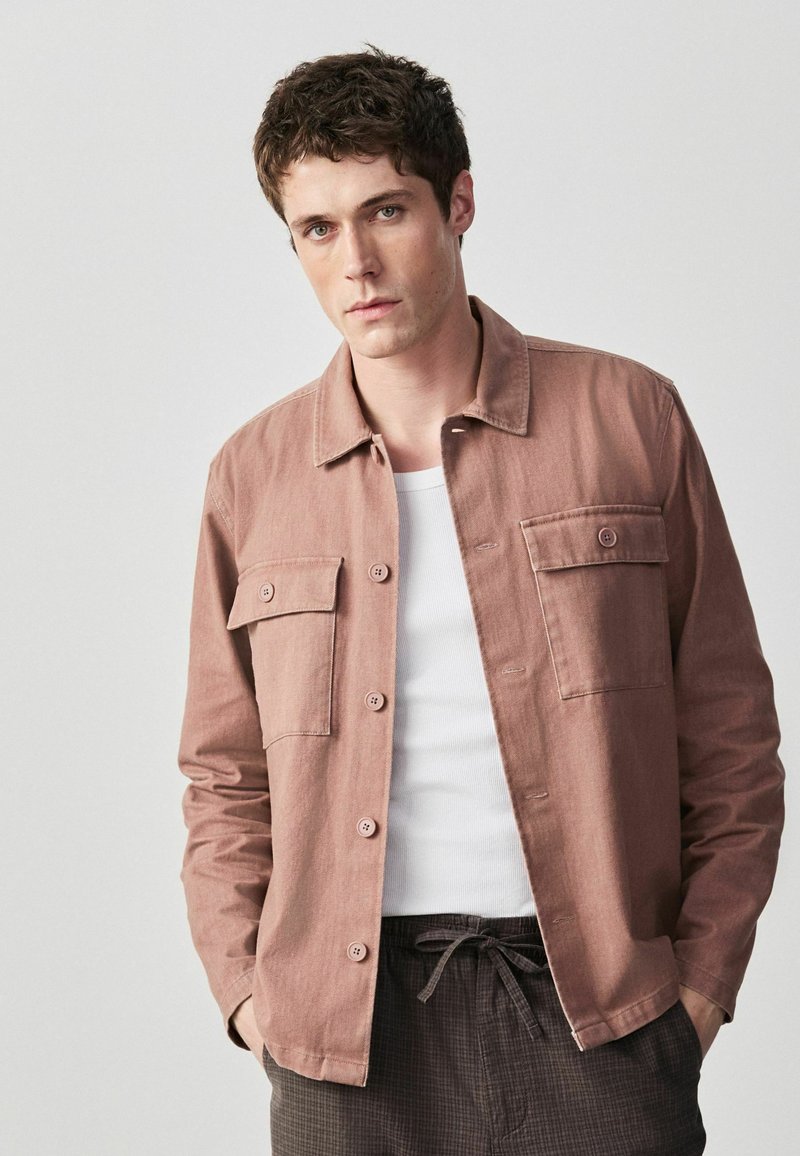 Jeune homme aux cheveux bruns courts portant une veste marron clair boutonnée sur une chemise blanche, debout les mains dans les poches d'un pantalon foncé.