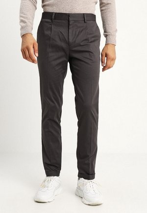Homme portant un pantalon slim foncé, un pull beige et des baskets blanches, debout devant un fond uni.