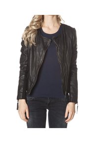 Femme portant une veste en cuir noire sur une chemise bleu marine et un jean foncé, debout devant un fond blanc.