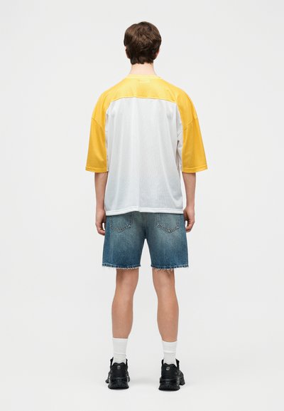 Άνδρας με φαρδιά λευκή και κίτρινη κοντομάνικη μπλούζα, σκισμένο τζιν σορτς, λευκές κάλτσες και μαύρα αθλητικά παπούτσια, στέκεται γυρισμένος με την πλάτη.