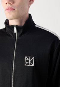 Zwarte zip-up jack van gladde stof, met een hoge kraag, contrasterende witte strepen op de schouders en een geborduurd logo.