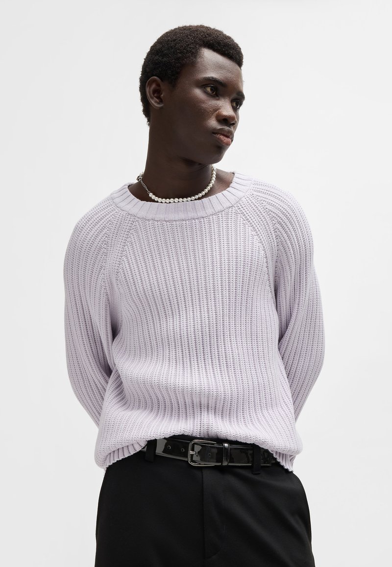 Pull en maille côtelée lavande clair avec un décolleté large et des manches raglan, porté avec une ceinture noire, sur un fond blanc uni.