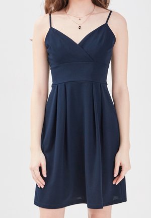 Woman wearing sleeveless dark blue V-neck dress with thin straps and pleated skirt, shown from shoulders to mid-thigh.