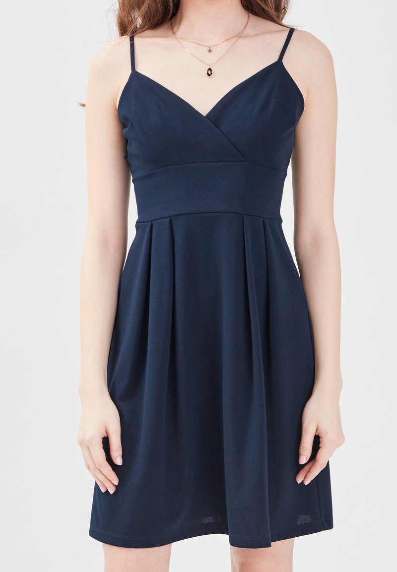 Femme portant une robe bleue foncée sans manches à encolure en V avec fines bretelles et jupe plissée, montrée des épaules jusqu'au milieu de la cuisse.