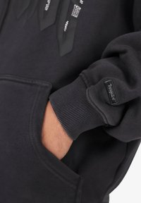 Hånd i lommen på en sort hættetrøje, der viser ribbede manchetter med et lille rektangulært sort mærke på ærmet.