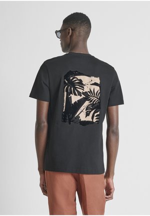 Hombre vistiendo camiseta negra con estampado de hojas tropicales en beige y negro en la espalda, combinada con pantalones de color óxido, de espaldas a la cámara.