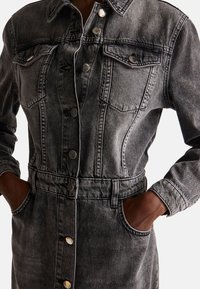 Abito in denim grigio con due tasche sul petto e vita con cintura. Presenta bottoni in metallo e dettagli cuciti, con maniche lunghe e tasche laterali.