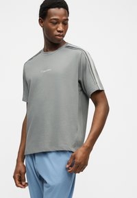 Άνδρας που φορά γκρι μπλουζάκι Calvin Klein με ρίγες στους ώμους και ανοιχτό μπλε παντελόνι, κοιτάζει προς τα κάτω δεξιά του.